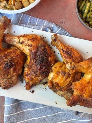 Roasted Chicken Leg Quarters placed on a white serving tray with vegetable sides in the background.