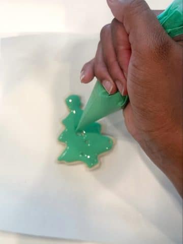 A hand decorating a Christmas tree-shaped cookie with green icing.
