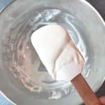 A spatula covered in homemade white royal icing, being held over a mixing bowl.
