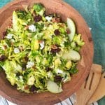A wooden bowl containing shaved Brussels sprouts salad with two wooden utensils next to the bowl.