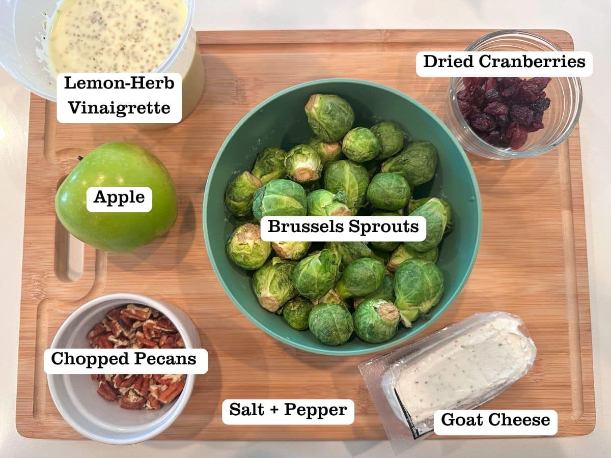 A wooden board containing bowls of brussels sprouts, pecans, dried cranberries, and a whole apple.