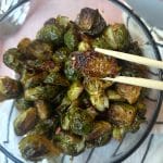 A chopstick holding up a roasted balsamic brussels sprout over a full bowl.