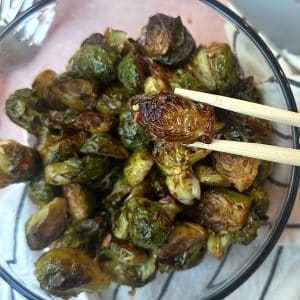 A chopstick holding up a roasted balsamic brussels sprout over a full bowl.