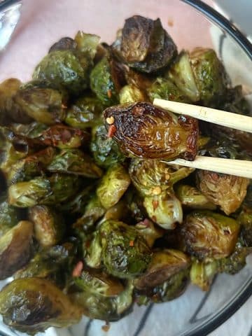 A chopstick holding up a roasted balsamic brussels sprout over a full bowl.
