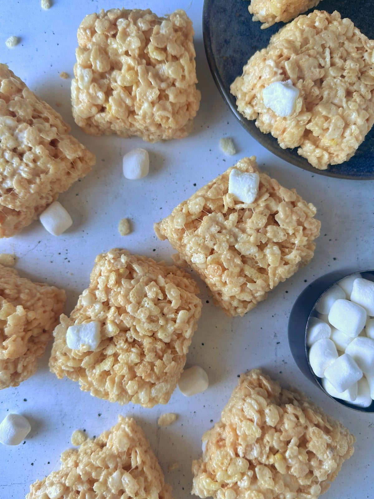 Multiple homemade cereal bar treats laid on a counter with extra marshmallows on the side.