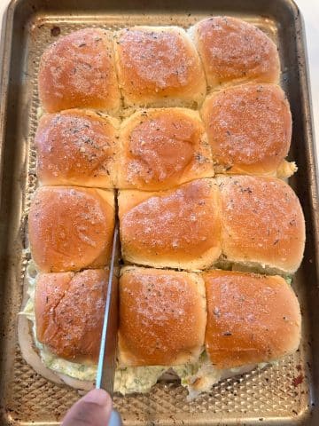 A knife slicing a block of meat and cheese stuffed rolls into individual sliders.