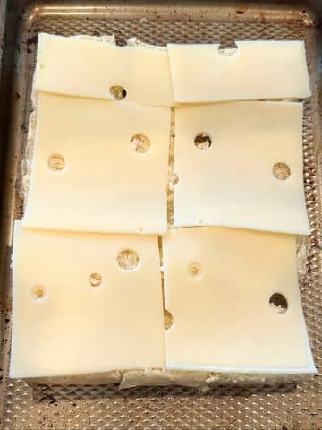 A sheet pan topped with Swiss cheese slices on a block of bottom buns.