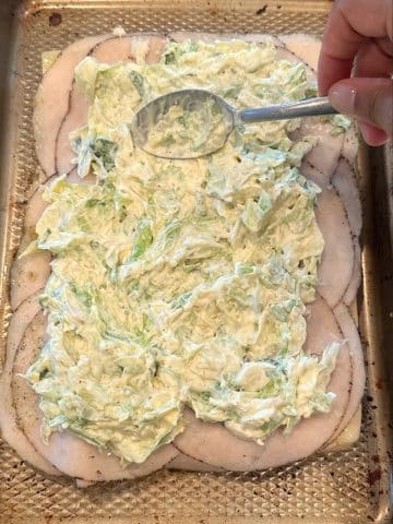 A hand using a spoon to spread coleslaw onto halved bread rolls topped with cheese and deli meat.