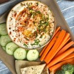 A bowl of whipped feta dip dressed with garnishments and served alongside cut vegetables and sliced pita.