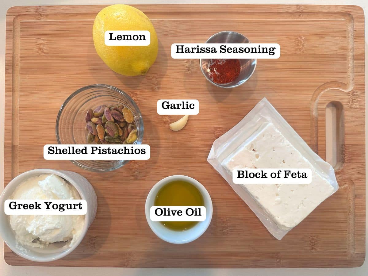 A wooden board filled with ingredients for whipped feta dip, including feta, harissa, and pistachios.
