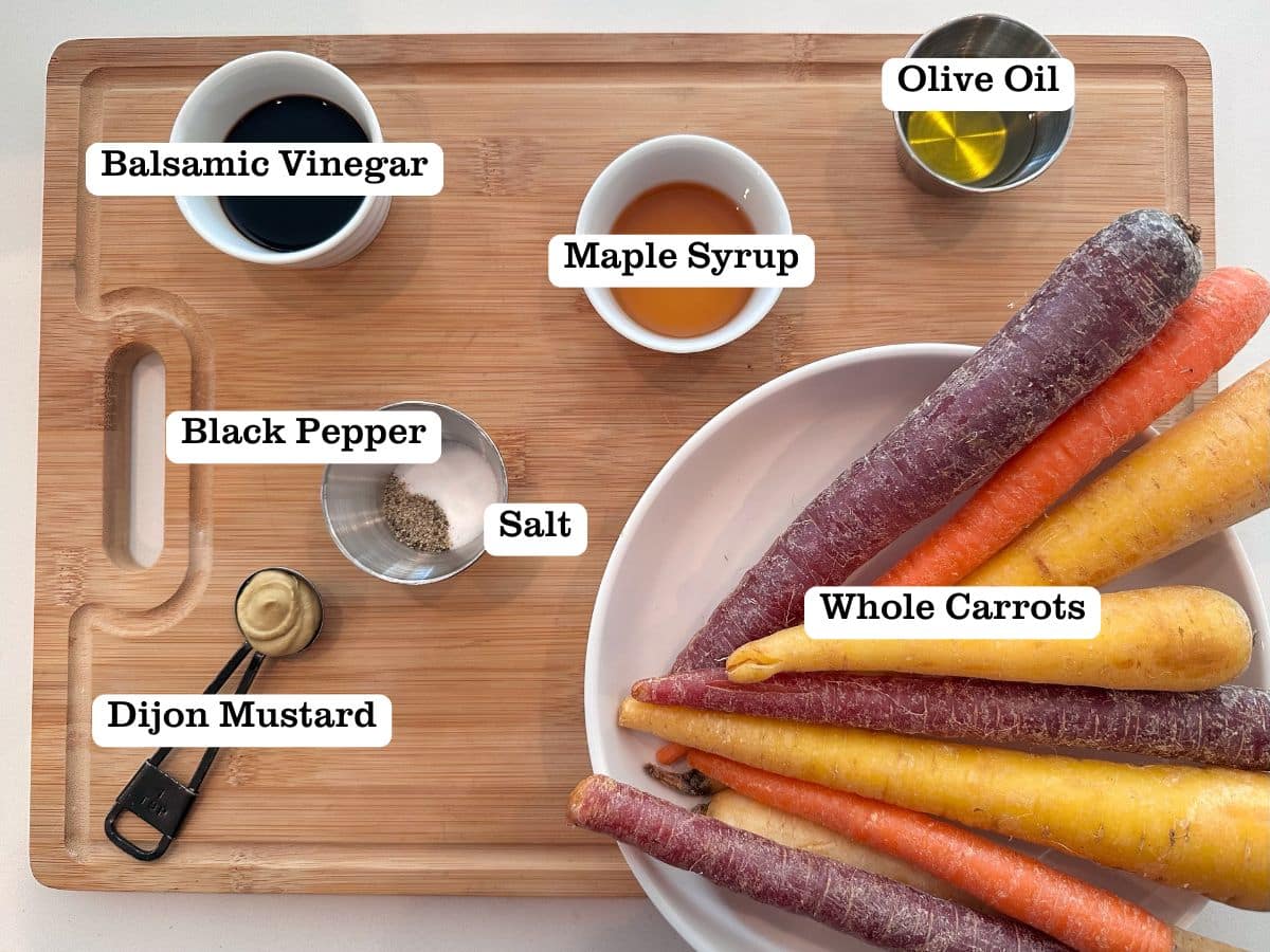 A wooden board containing ingredients for roasted carrots, including bowls of maple syrup, balsamic vinegar, and dijon mustard.