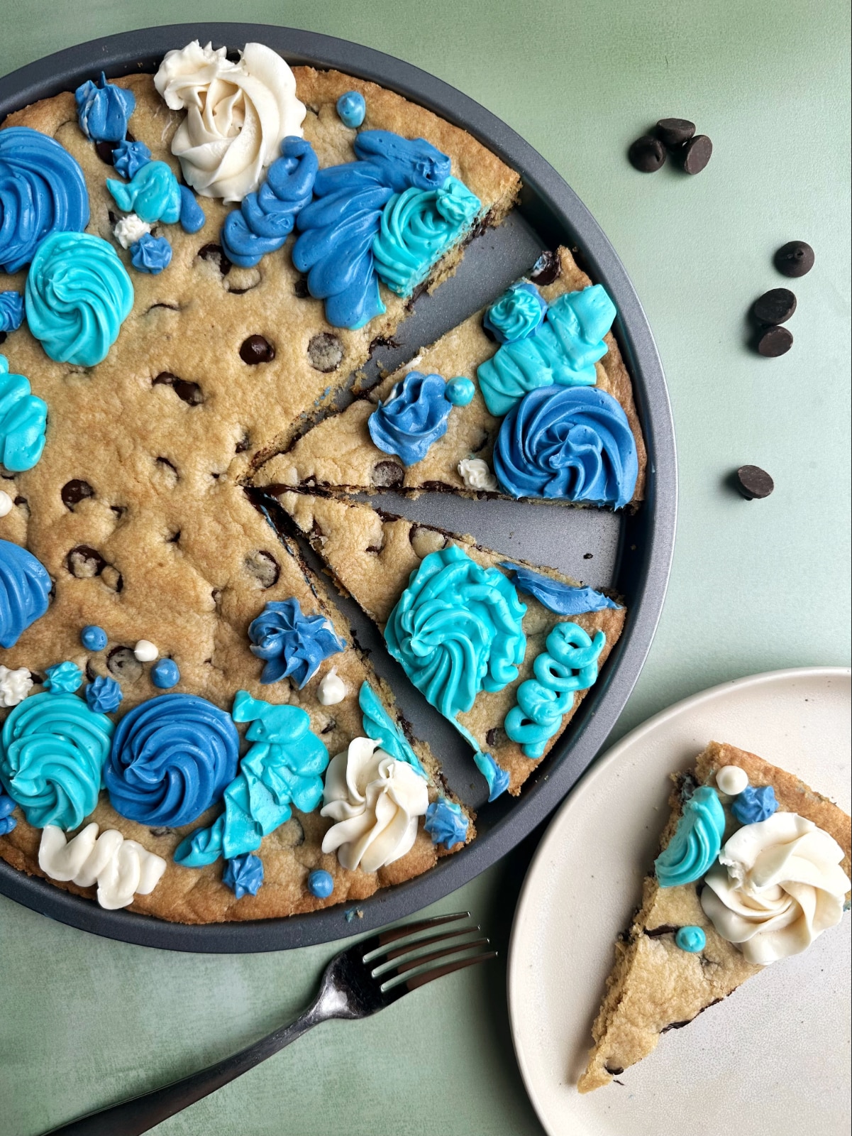 A cookie cake decorated with blue and white buttercream, with one slice cut out onto a plate.