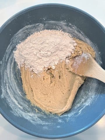 A wooden spoon stirring flour into a light brown cookie dough.