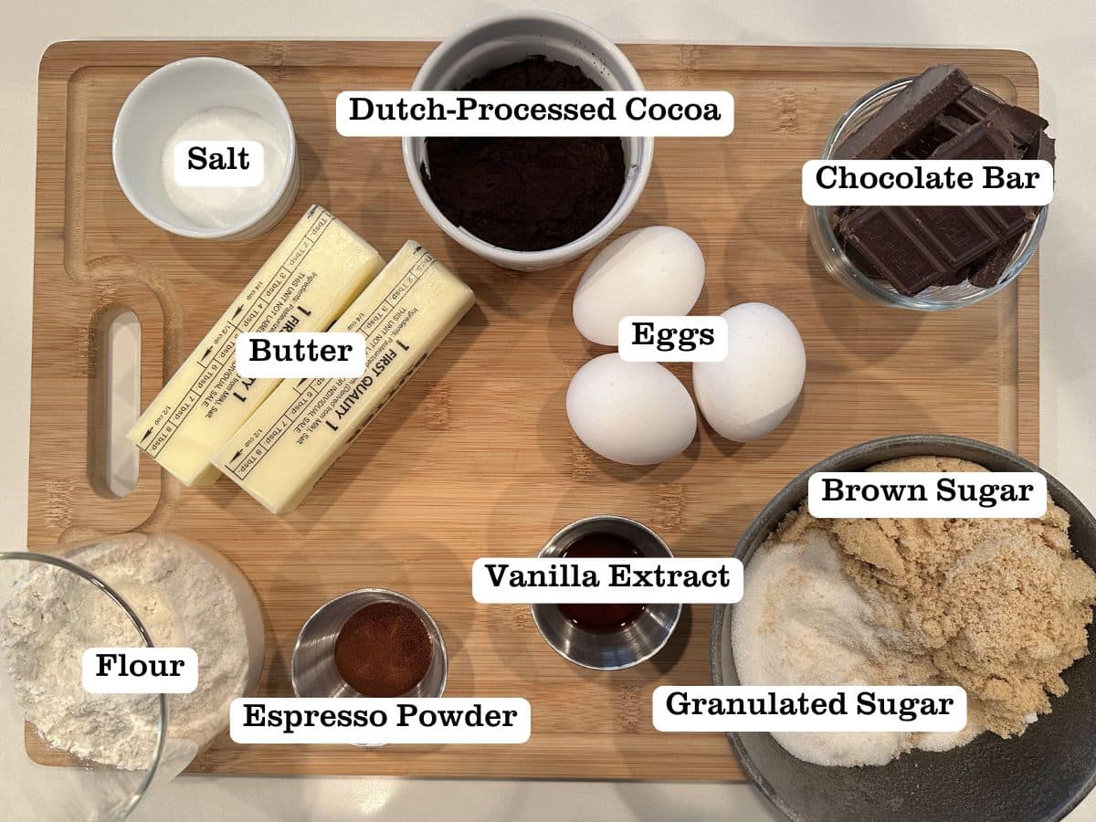 A wooden board containing ingredients for crinkle top fudge brownies, including butter, bowls of cocoa powder, espresso powder, and brown sugar.