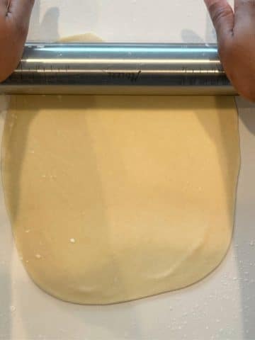 Two hands using a silver rolling pin to roll out tortilla dough into a thin disc.