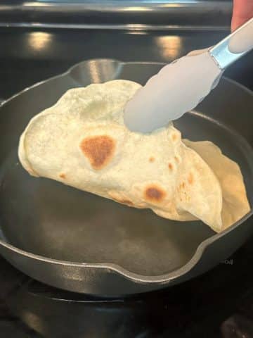 A hand using a pair of tongs to lift up a tortilla out of a cast iron skillet.