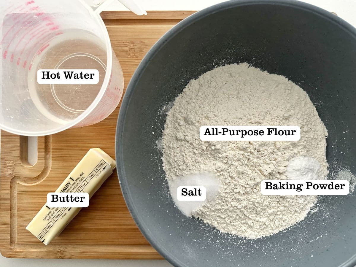A wooden board containing ingredients for flour tortillas from scratch, including a stick of butter and a bowl of hot water.