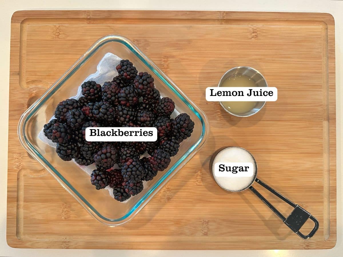 Containers of fresh blackberries, white sugar, and lemon juice on a wooden board.