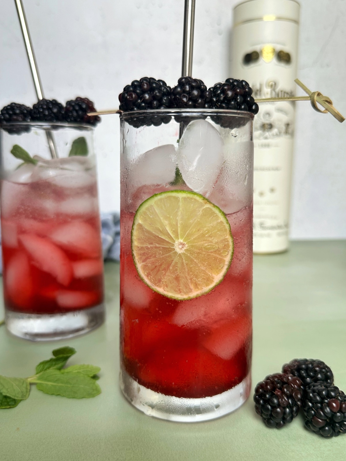 Two glasses of blackberry midnight mojitos served over ice, with a bottle of rum in the background.