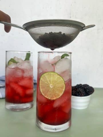 Muddled blackberries being strained over a glass of club soda.