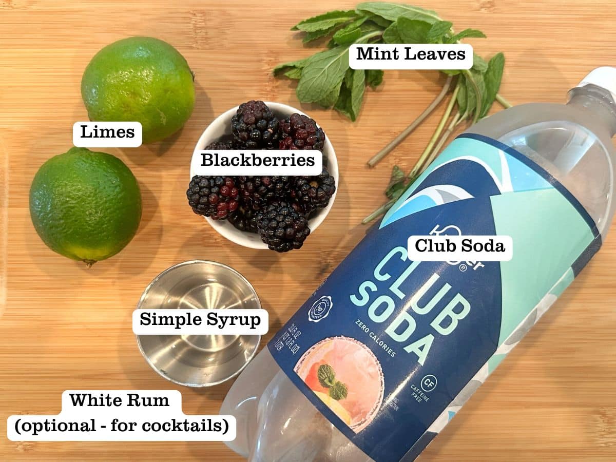 A wooden board containing ingredients for blackberry mojitos, including club soda, cups of blackberries, and simple syrup.