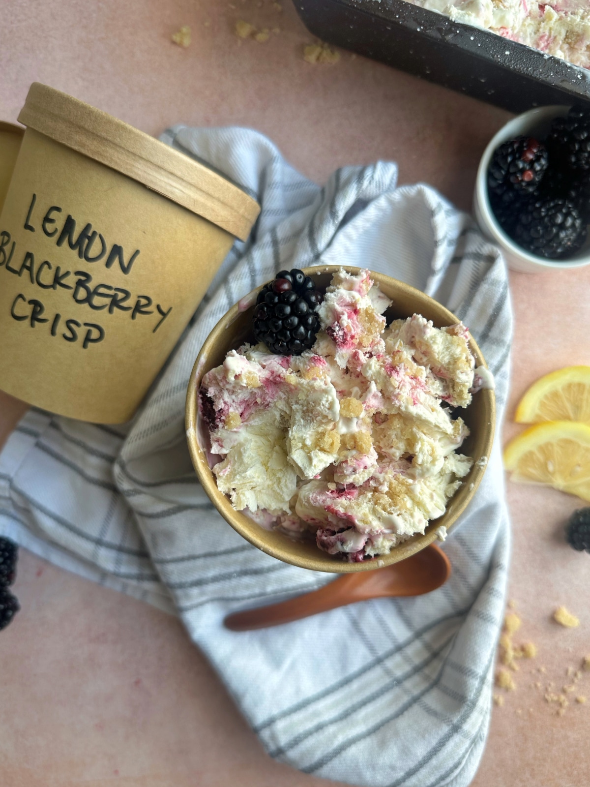 A brown ice cream container filled with ice cream and blackberries, with another labeled container on the side that says "Lemon Blackberry Crisp".