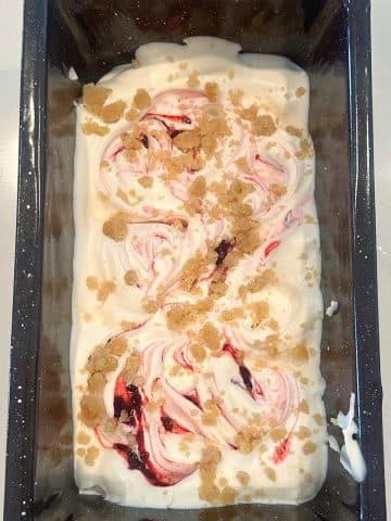 A loaf pan filled with lemon ice cream with blackberry swirls and crumbled cookies on top.