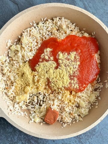 A skillet filled with toasted rice, onions, tomato sauce, and yellow and red seasonings.