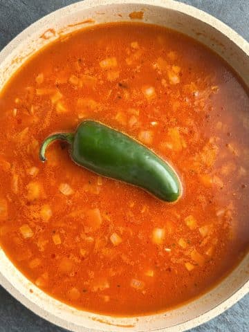A tomato-based, soupy liquid with rice and a whole jalapeño in a skillet.