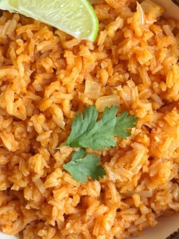 A bowl of Mexican red rice with a lime quarter and a few sprigs of cilantro.