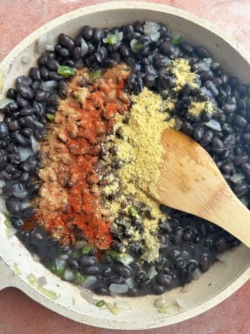 A pan of black beans with red and yellow dried seasonings on top, and a wooden spoon sticking out.