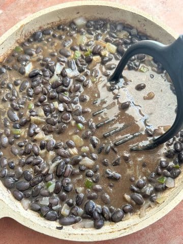 A pan of black beans being mashed with a. potato masher utensil.