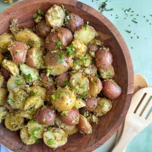 A wooden bowl containing Chimichurri potato salad with serving utensils on the side.