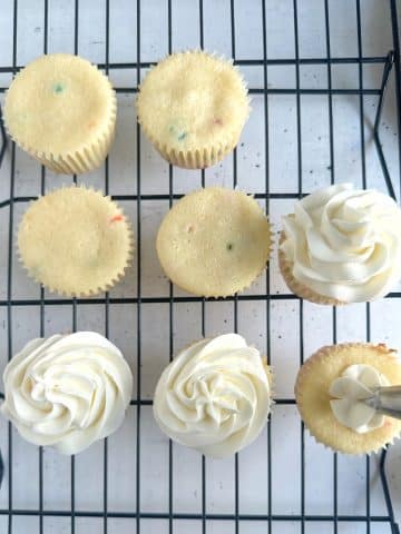 Vanilla cupcakes on a wire rack being frosted with vanilla buttercream.
