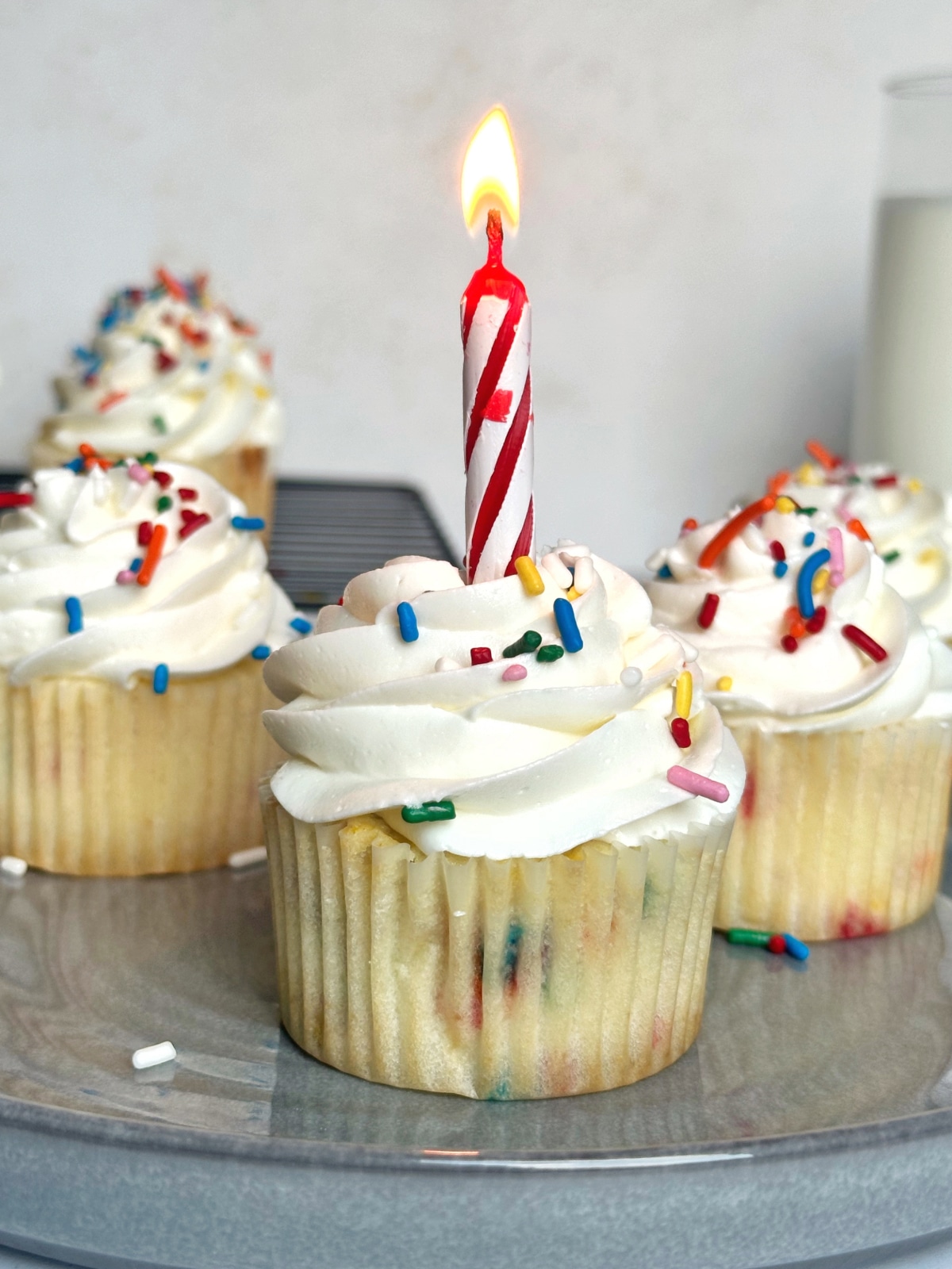 A plate of cupcakes with rainbow sprinkles and a lit birthday candle in the center of the first cupcake.