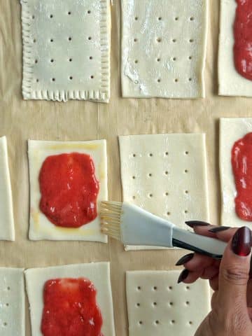 Egg wash being brushed on the edge of a pastry sheet with red filling in the middle.