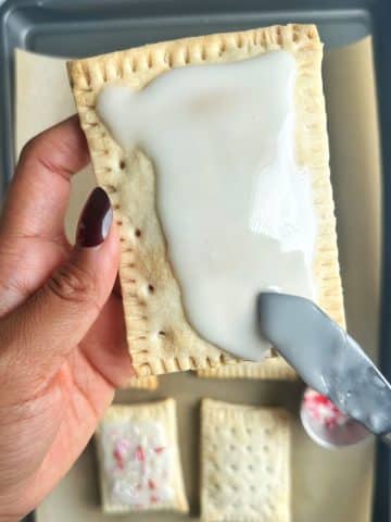 A hand holding a homemade pop tart, spreading white icing on it with an offset spatula.