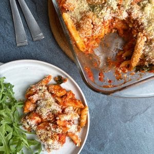 A plate of chicken parmesan pasta bake next to the pan with the rest of the dish inside.