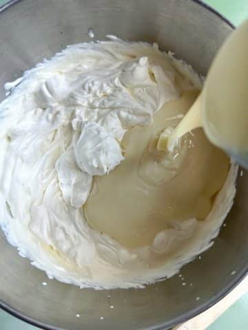 A mixing bowl with whipped cream and an off-camera hand pouring a can of sweetened condensed milk inside.