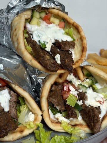 A stack of three lamb gyro wraps with fries on a plate in the background.