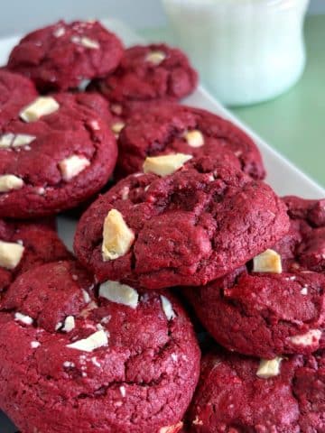 A stack of red velvet cake mix cookies on a plate with a glass of milk in the background.