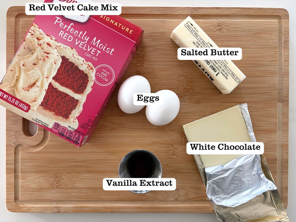 A wooden board containing ingredients for red velvet cake mix cookies, including white chocolate and eggs.