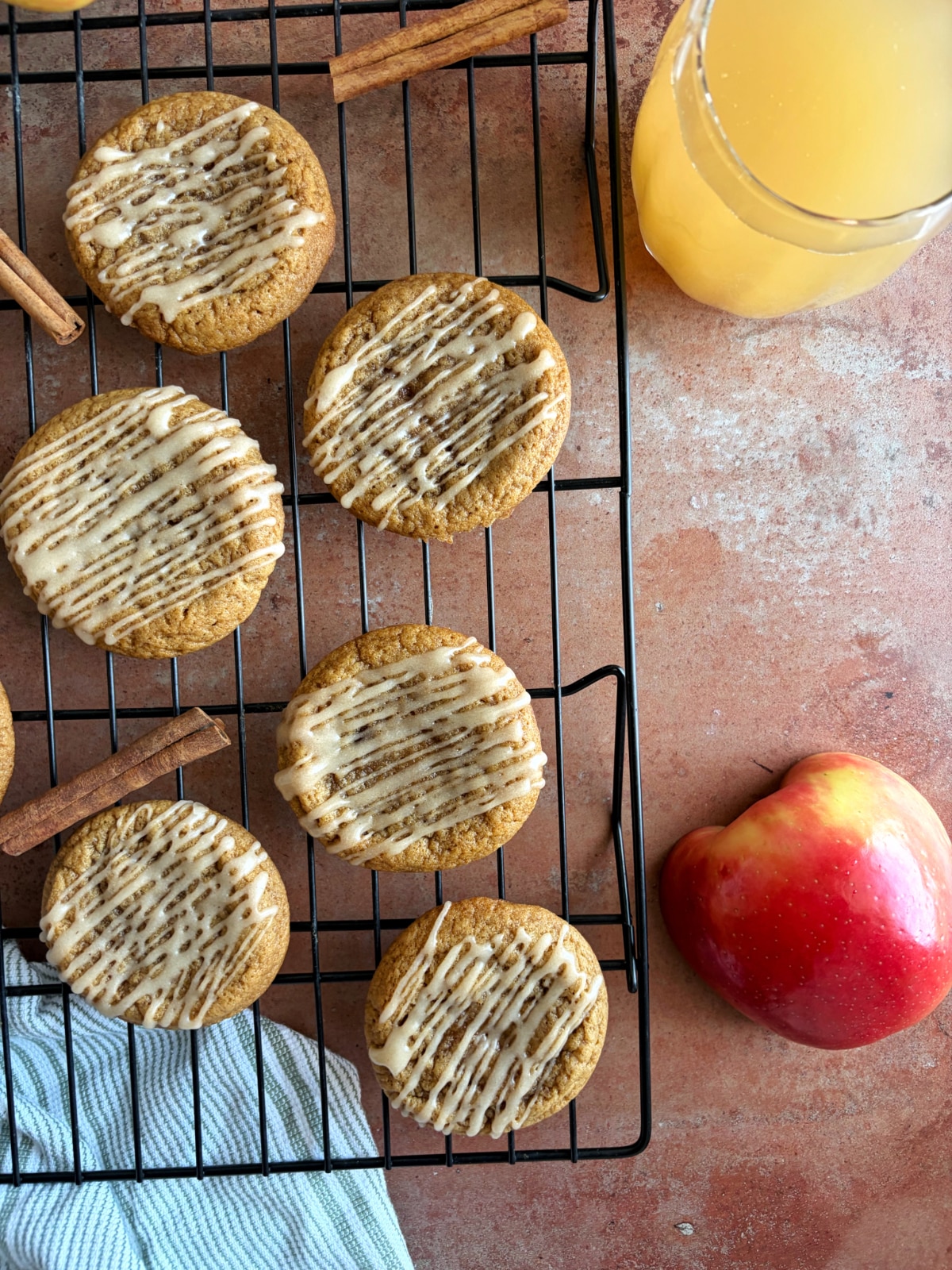 A wire rack filled with drizzled cookies, with a glass of apple cider and cinnamon sticks on the side.