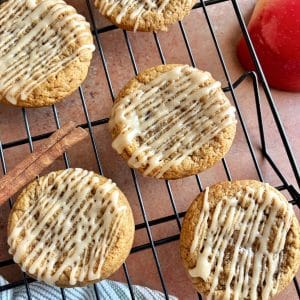 Apple Cider Cookies with a cider drizzle placed on a wire cooling rack, with cinnamon sticks and apple halves in the background.