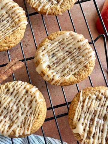 Apple Cider Cookies with a cider drizzle placed on a wire cooling rack, with cinnamon sticks and apple halves in the background.