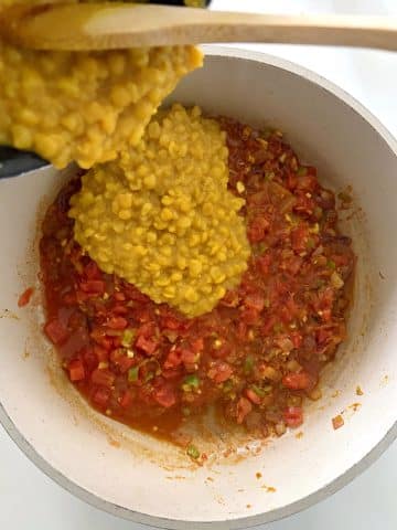 Cooked yellow lentils being poured into a skillet with stewed tomatoes and onions.