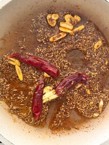 Dried red chilies, whole spices, and sliced garlic sautéed in ghee in a skillet.