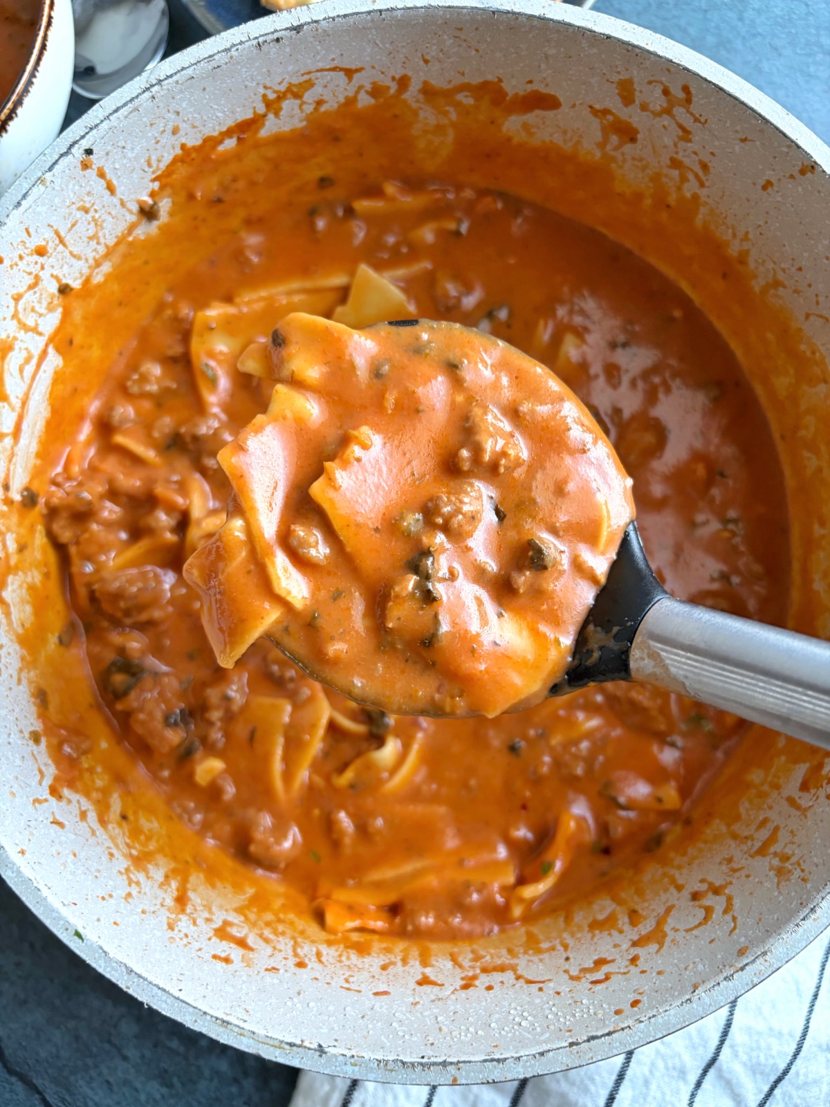A ladle of red sauce soup with pasta, held over the rest of the batch in a cream-colored pot.