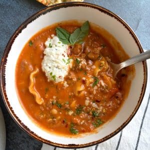 A bowl of lasagna soup with a dollop of ricotta cheese, basil, and fresh herbs as garnish.