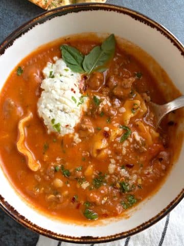 A bowl of lasagna soup with a dollop of ricotta cheese, basil, and fresh herbs as garnish.